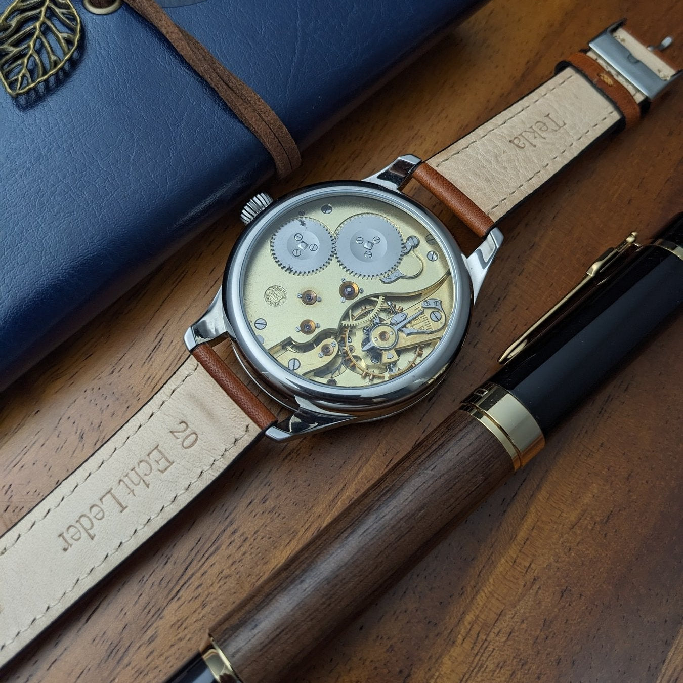 Watch with brown leather strap on a wooden surface next to a blue notebook and pen.